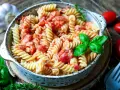 Fusilli-Nudeln in cremiger Tomaten-Sahne-Sauce, garniert mit frischem Basilikum. Daneben liegen Kirschtomaten und Knoblauch.