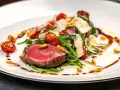 Ein saftiges Steak, italienisch zubereitet, mit Rucola, Kirschtomaten, Pinienkernen und Parmesanspänen auf einem weißen Teller.