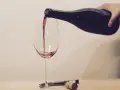Ein Weinglas wird mit Rotwein gefüllt. Eine Hand hält die dunkle Flasche, während der Wein in einem dünnen Strahl ins Glas fließt. Ein Korkenzieher und ein Korken liegen daneben.