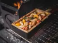 Salsiccia grillen in einer Keramikform mit Tomaten und Kräutern. Flammen lodern im Hintergrund.