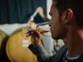 Mann verkostet bernsteinfarbenen italienischen Dessertwein in einem Glas, im Hintergrund Weinfässer.