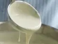 Cremige Bechamelsoße tropft von einer Schöpfkelle in einen Topf. Perfekt für Lasagne!