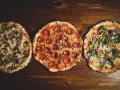 Drei italienische Pizzen: eine mit Pilzen, eine mit Salami und eine mit Gemüse, liegen auf einem Holztisch.