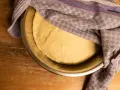 Pizzateig ruht in einer Schüssel, bedeckt mit einem gemusterten Tuch. Die Frage "wie lange pizzateig gehen lassen" beschäftigt den Bäcker.