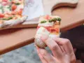 Eine Hand hält ein Stück Pizza mit Tomaten, Mozzarella und Pesto. So sieht eine köstliche Pizza aus!