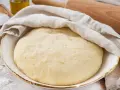 Frisch zubereiteter pizzateig mit trockenhefe ruht unter einem Tuch. Daneben liegen Salz und Olivenöl.