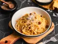 Eine Schüssel mit echter Carbonara, garniert mit geriebenem Parmesan und schwarzem Pfeffer. Daneben ein Holzlöffel mit Pfefferkörnern.