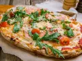 Eine köstliche Pinsa belegt mit Schinken, Rucola, Kirschtomaten und viel Käse auf rustikalem Holzuntergrund.