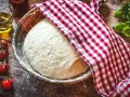 Frischer Pizzateig ruht in einer Schüssel, bedeckt mit einem rot-weiß karierten Tuch. Daneben liegen Kirschtomaten und ein Glas Olivenöl – die perfekten pizzateig zutaten.