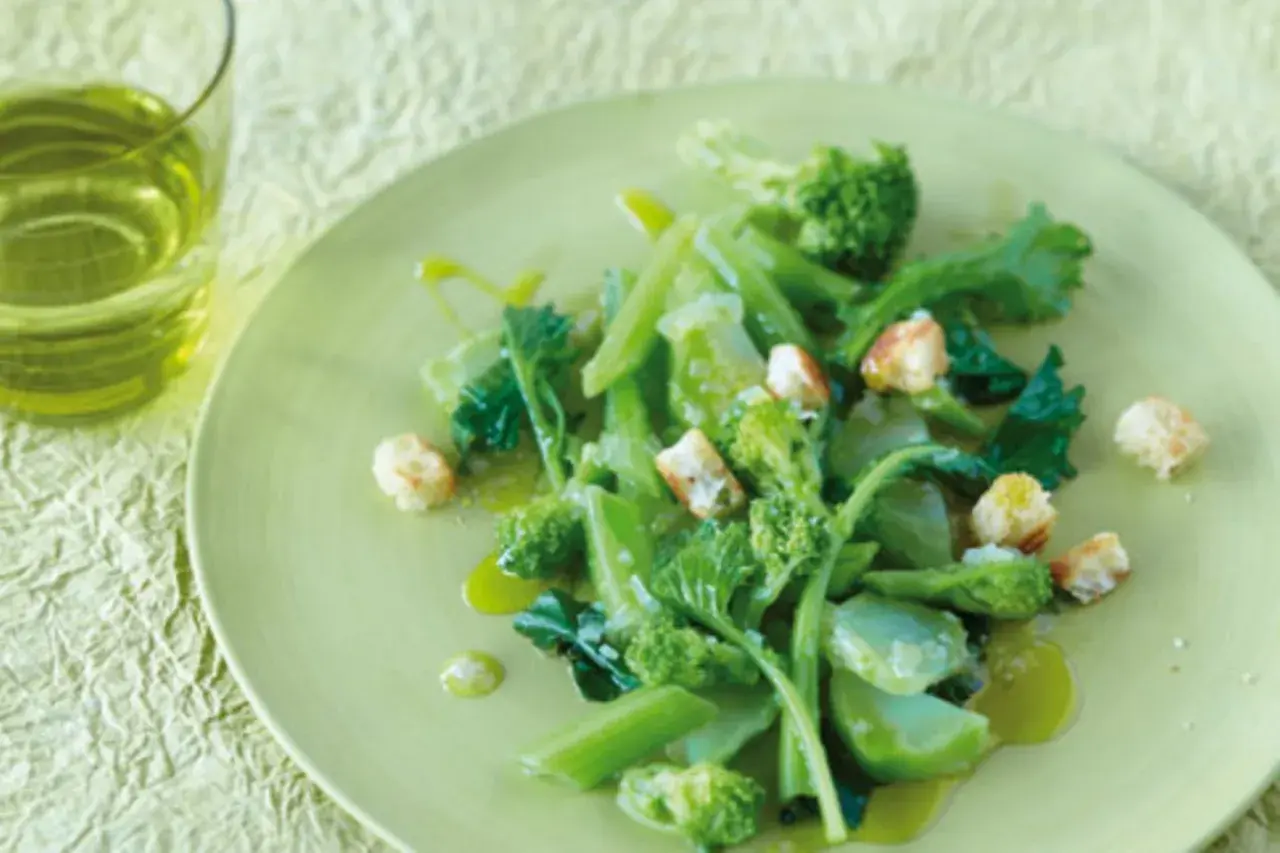 Ein Teller mit einem frischen Salat aus wildem Brokkoli, garniert mit Croutons und beträufelt mit Olivenöl. Daneben ein Glas grünes Getränk.