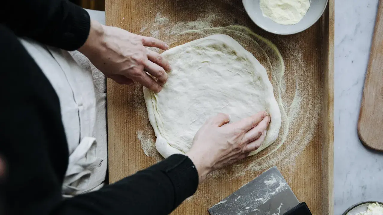 Hände formen einen Teig für ein Pizzateig Rezept auf einem Holzbrett. Mehl und ein Teigschaber liegen bereit.
