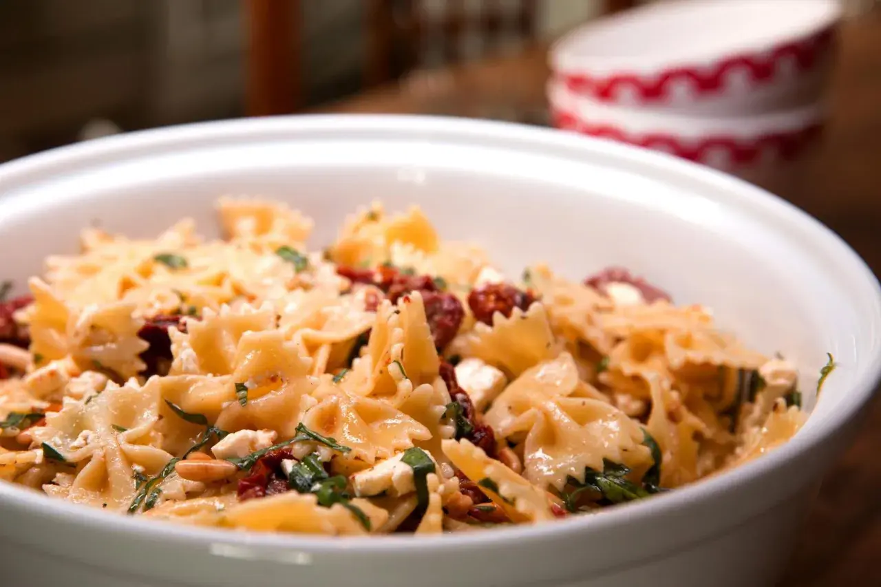 Ein köstlicher italienischer Nudelsalat mit Farfalle, getrockneten Tomaten, Feta und Kräutern in einer weißen Schüssel.