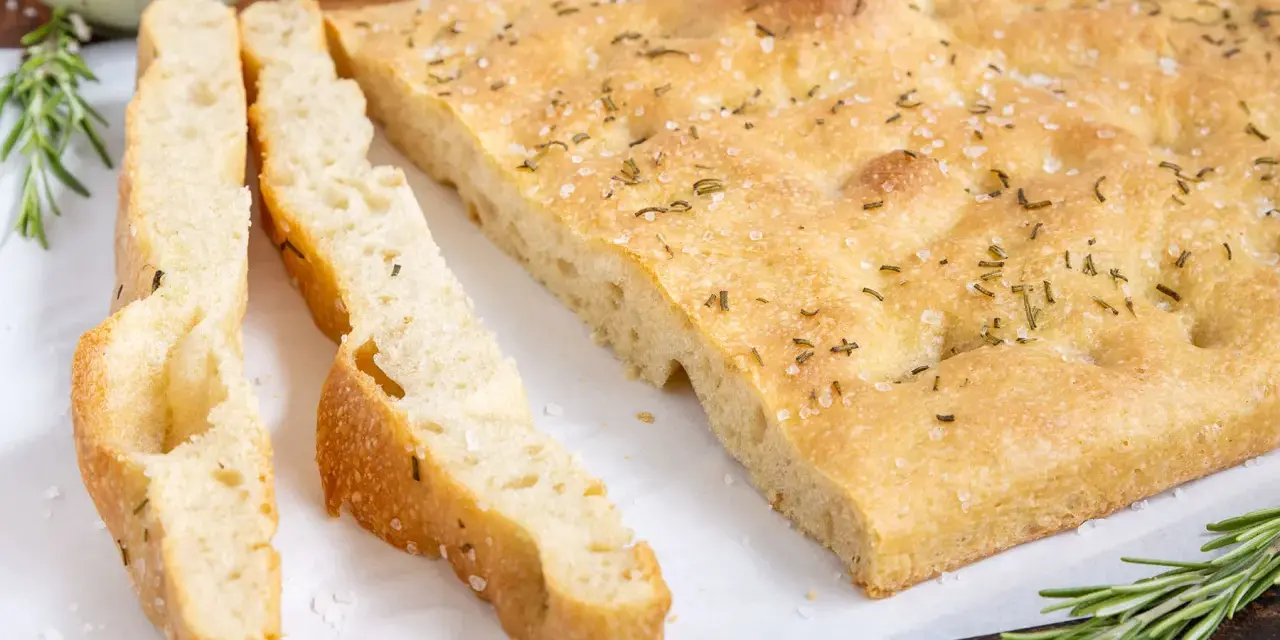 Frisch gebackene Focaccia, ein Klassiker unter den italienischen Brotsorten, mit grobem Salz und Rosmarin bestreut. Daneben liegen zwei geschnittene Stücke.