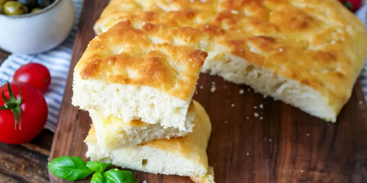 Frisch gebackener focaccia teig, aufgeschnitten und gestapelt, mit Tomaten und Oliven im Hintergrund.