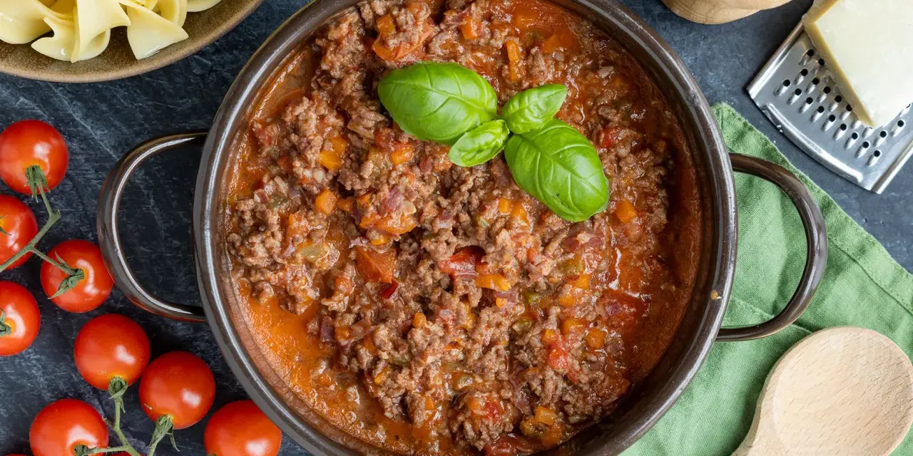 Herzhaftes ragù alla bolognese in einem Topf, garniert mit Basilikum. Daneben frische Tomaten, Bandnudeln und Parmesan.