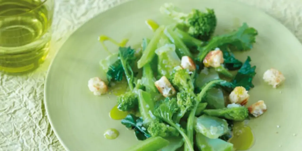 Ein Teller mit einem frischen Salat aus wildem Brokkoli, garniert mit Croutons und beträufelt mit Olivenöl. Daneben ein Glas grünes Getränk.