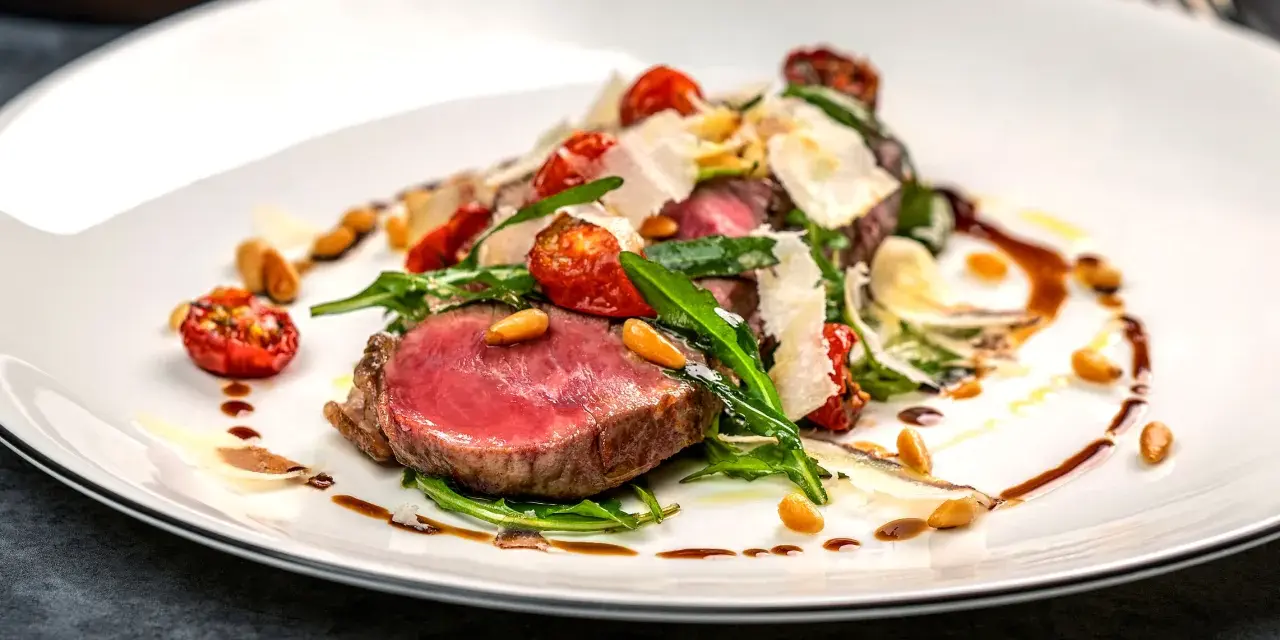 Ein saftiges Steak, italienisch zubereitet, mit Rucola, Kirschtomaten, Pinienkernen und Parmesanspänen auf einem weißen Teller.