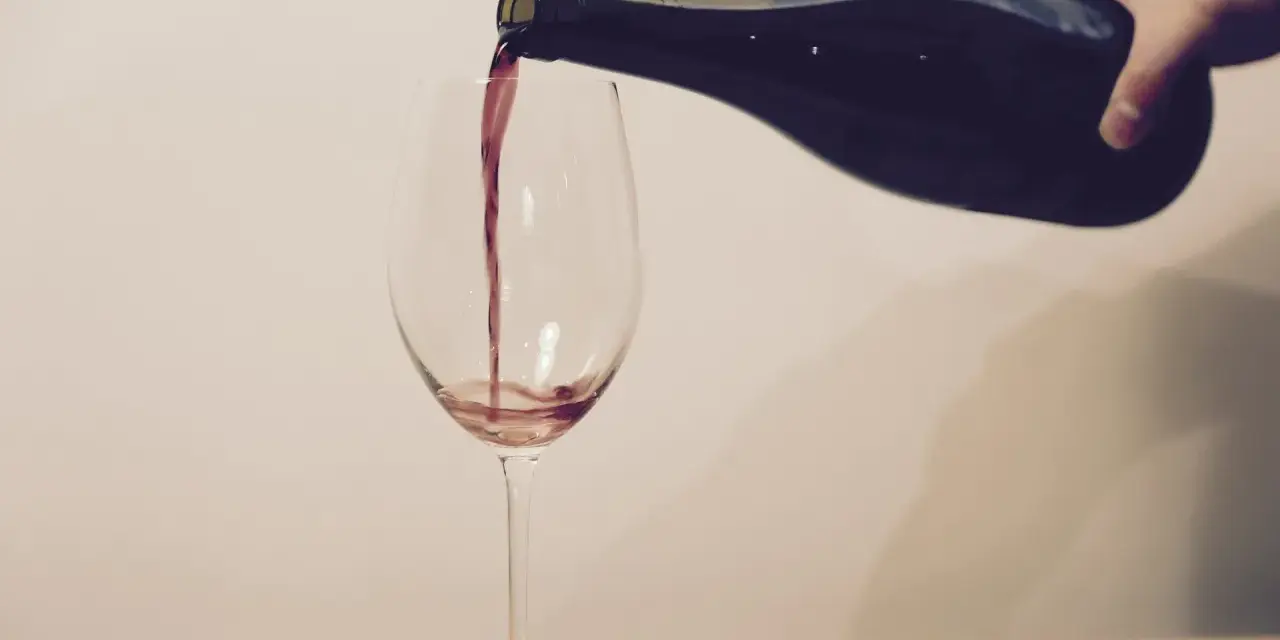 Ein Weinglas wird mit Rotwein gefüllt. Eine Hand hält die dunkle Flasche, während der Wein in einem dünnen Strahl ins Glas fließt. Ein Korkenzieher und ein Korken liegen daneben.