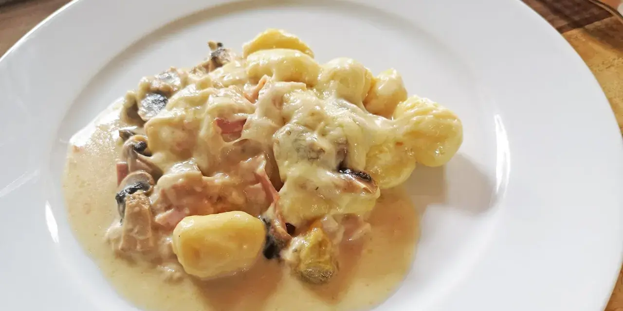 Gnocchi al forno mit cremiger Käsesauce, Pilzen und Schinken auf einem weißen Teller.