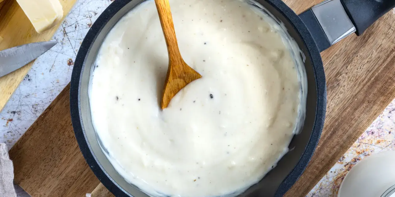 Cremige Béchamelsauce in einer Pfanne, bereit für Lasagne. Butter, Milch und Gewürze liegen daneben.