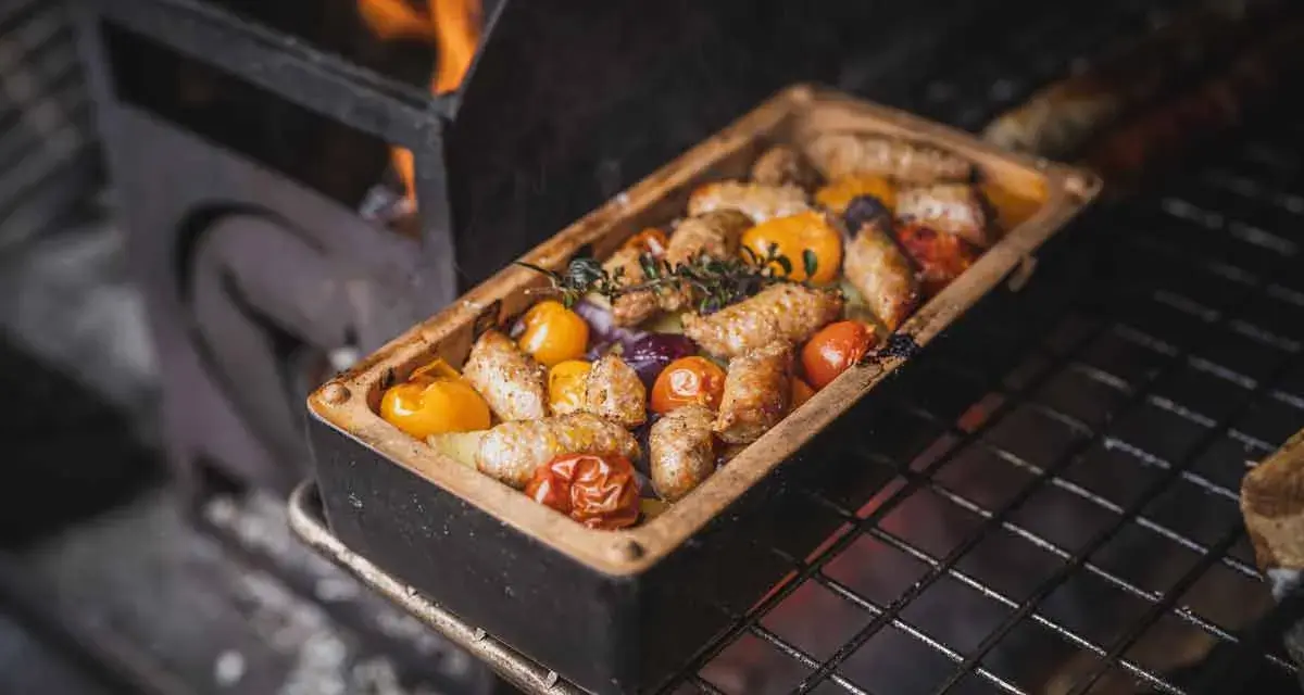 Salsiccia grillen in einer Keramikform mit Tomaten und Kräutern. Flammen lodern im Hintergrund.