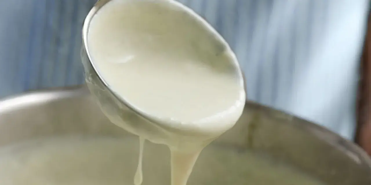 Cremige Bechamelsoße tropft von einer Schöpfkelle in einen Topf. Perfekt für Lasagne!