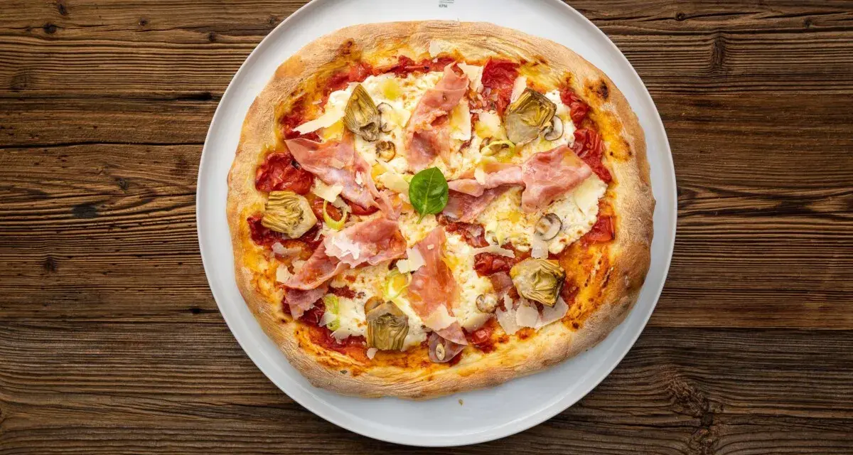Eine köstliche Prosciutto Pizza mit Artischocken und Basilikum auf einem weißen Teller, serviert auf rustikalem Holz.