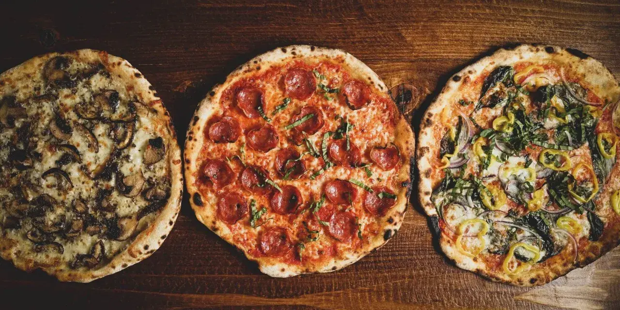 Drei italienische Pizzen: eine mit Pilzen, eine mit Salami und eine mit Gemüse, liegen auf einem Holztisch.