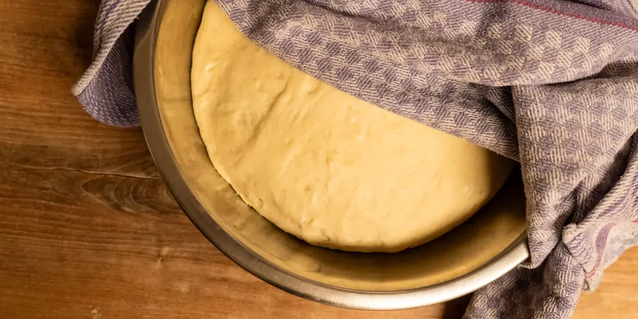 Pizzateig ruht in einer Schüssel, bedeckt mit einem gemusterten Tuch. Die Frage "wie lange pizzateig gehen lassen" beschäftigt den Bäcker.