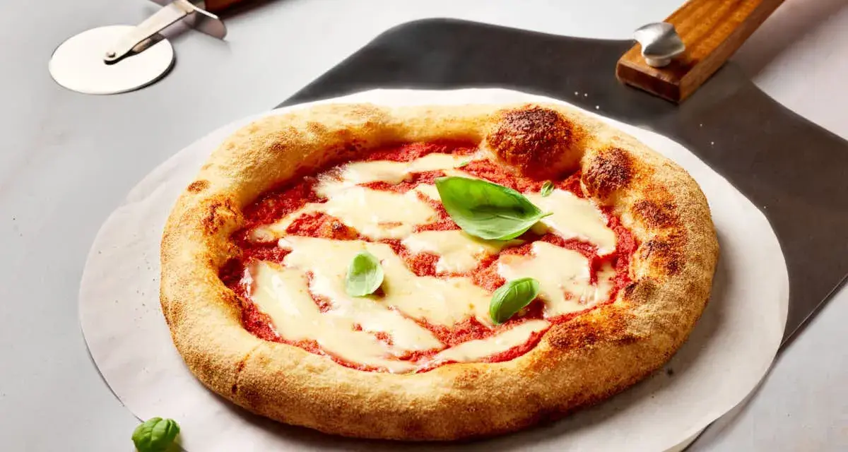 Frisch gebackene Pizza Margherita mit geschmolzenem Mozzarella und Basilikum. Daneben liegen ein Pizzaschneider und eine Pizzaschaufel, bereit, den perfekten pizzateig ausrollen.