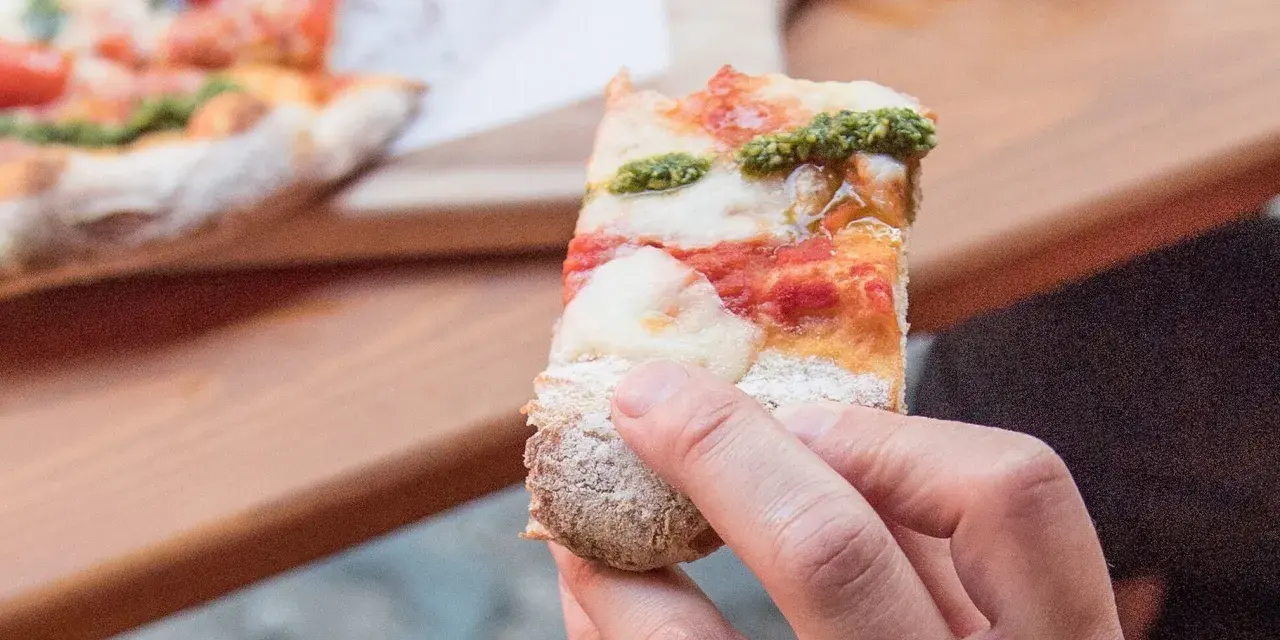 Eine Hand hält ein Stück Pizza mit Tomaten, Mozzarella und Pesto. So sieht eine köstliche Pizza aus!