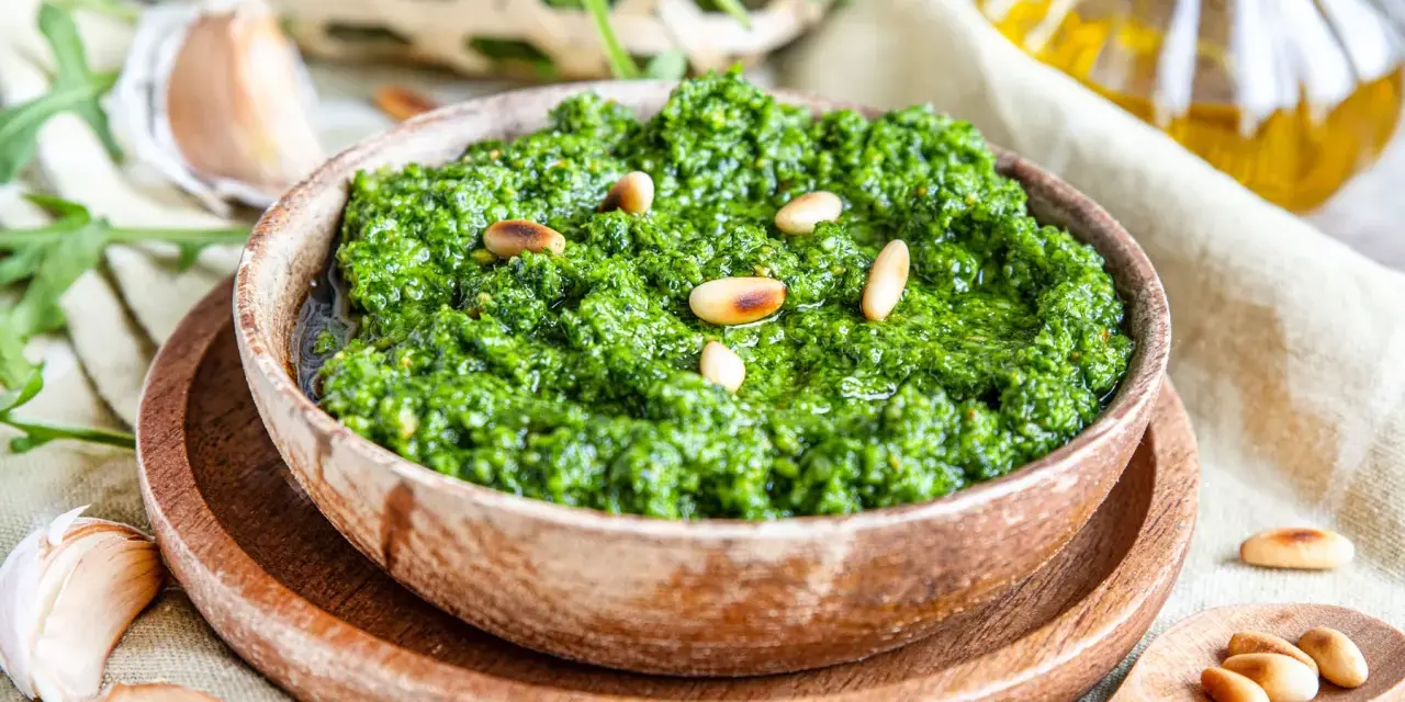 Frisches Rucola Pesto, garniert mit Pinienkernen, daneben Knoblauch und Olivenöl.
