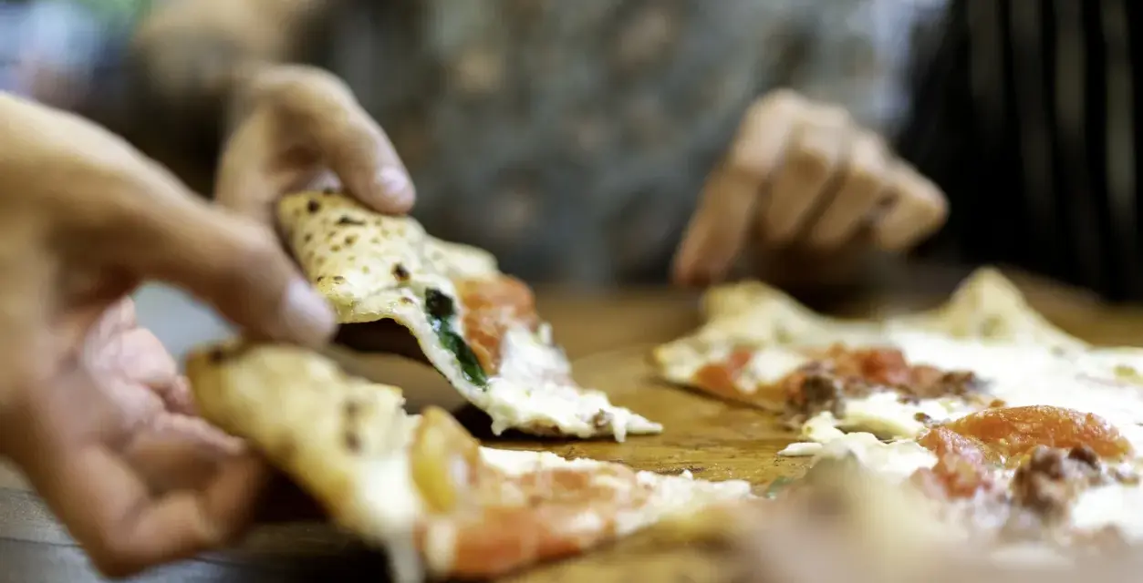 Hände greifen nach dampfenden Pizzastücken. So wie essen Italiener Pizza: gemeinsam, genussvoll, mit Freunden.