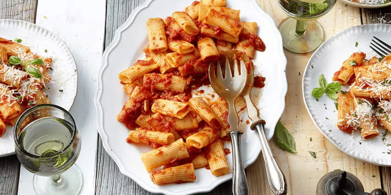 Ein Teller mit Rigatoni mit Tomatensauce, garniert mit Parmesan und Basilikum. Daneben ein Glas Weißwein.