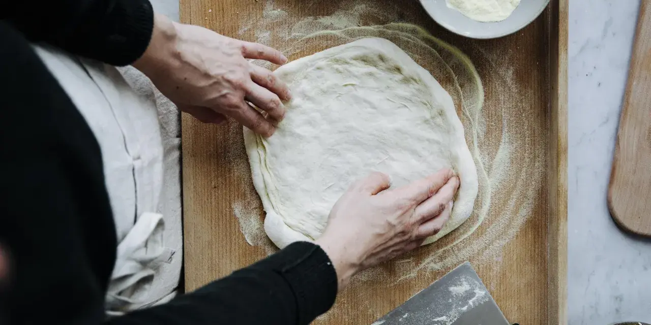 Hände formen einen Teig für ein Pizzateig Rezept auf einem Holzbrett. Mehl und ein Teigschaber liegen bereit.