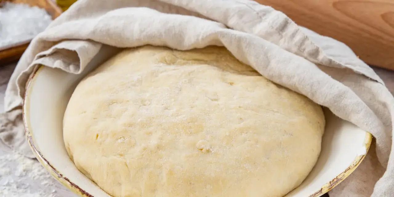 Frisch zubereiteter pizzateig mit trockenhefe ruht unter einem Tuch. Daneben liegen Salz und Olivenöl.