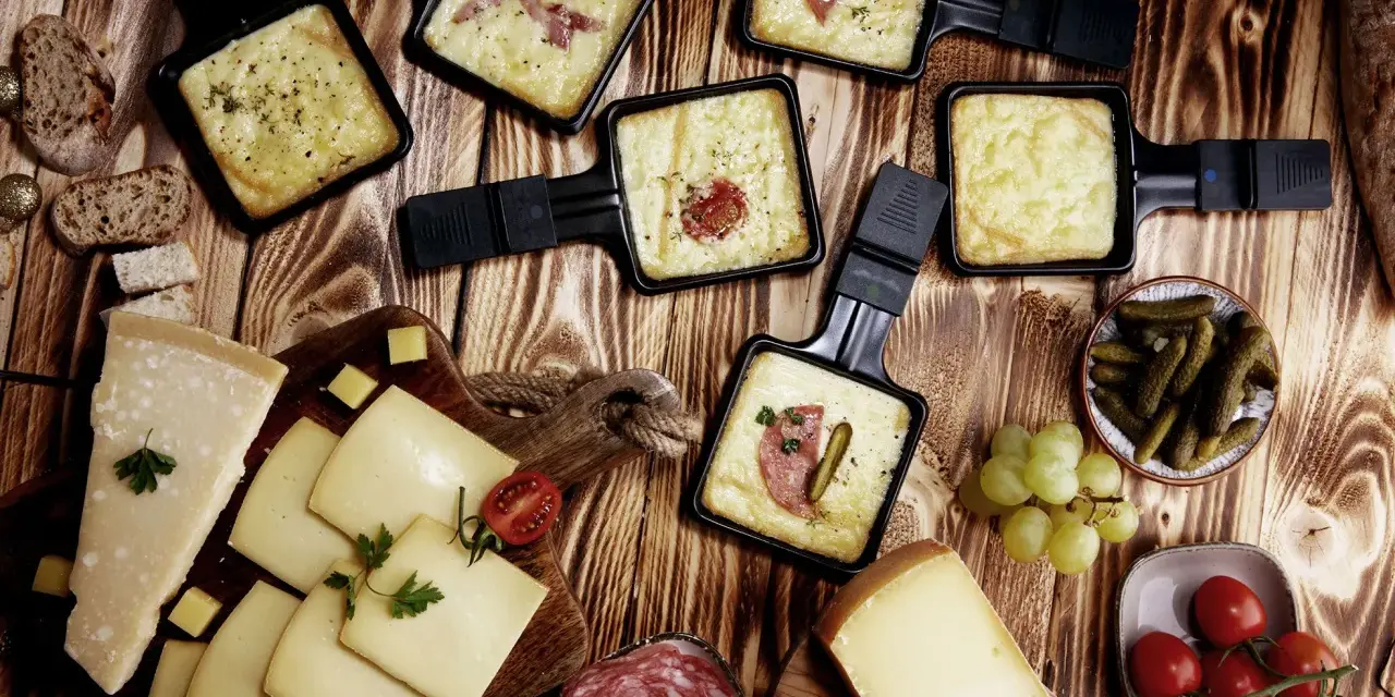 Raclette-Pfännchen mit geschmolzenem Käse, dazu Brot, Käse, Salami, Cornichons und Trauben. Perfekt für einen gemütlichen Abend mit **Wein zum Raclette**.