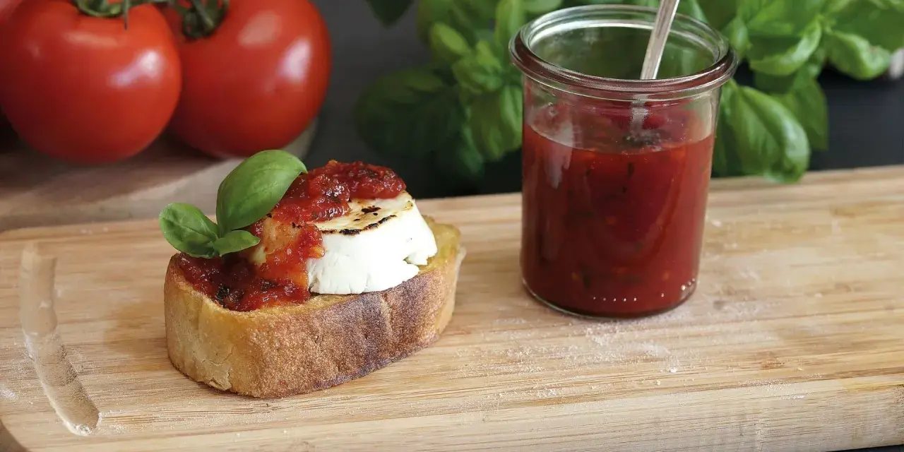 Bruschetta mit Ziegenkäse und Tomatenmarmelade, inspiriert von einem italienischen Tomatenmarmelade Rezept. Frische Tomaten und Basilikum im Hintergrund.