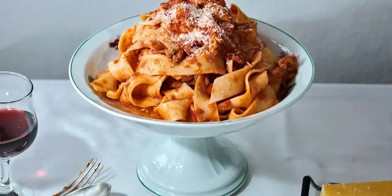 Ein Teller breiter Bandnudeln mit herzhaftem al ragu, bestreut mit Parmesan. Daneben ein Glas Rotwein und Besteck.