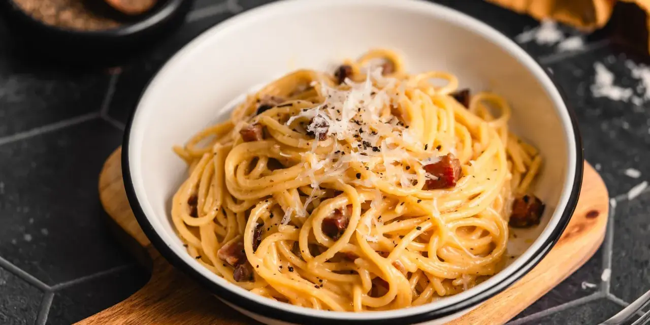 Eine Schüssel mit echter Carbonara, garniert mit geriebenem Parmesan und schwarzem Pfeffer. Daneben ein Holzlöffel mit Pfefferkörnern.