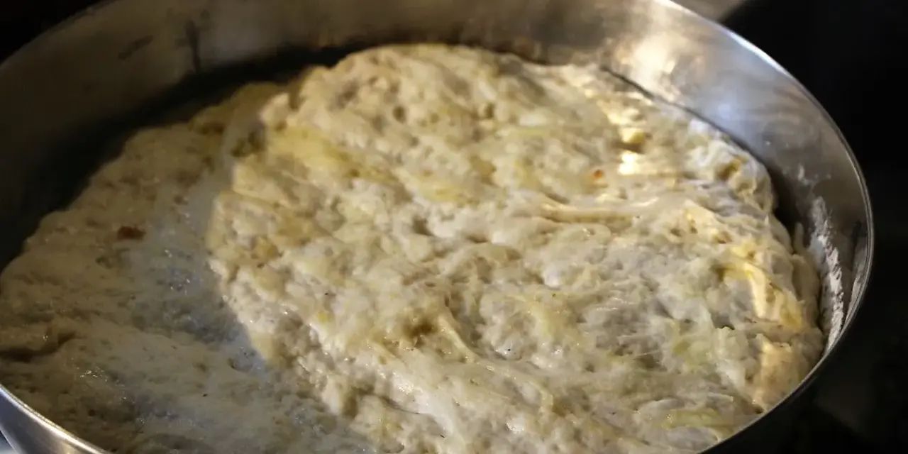 Hefeteig für Pizza geht auf. Die luftige Masse quillt über den Rand der Metallschüssel.