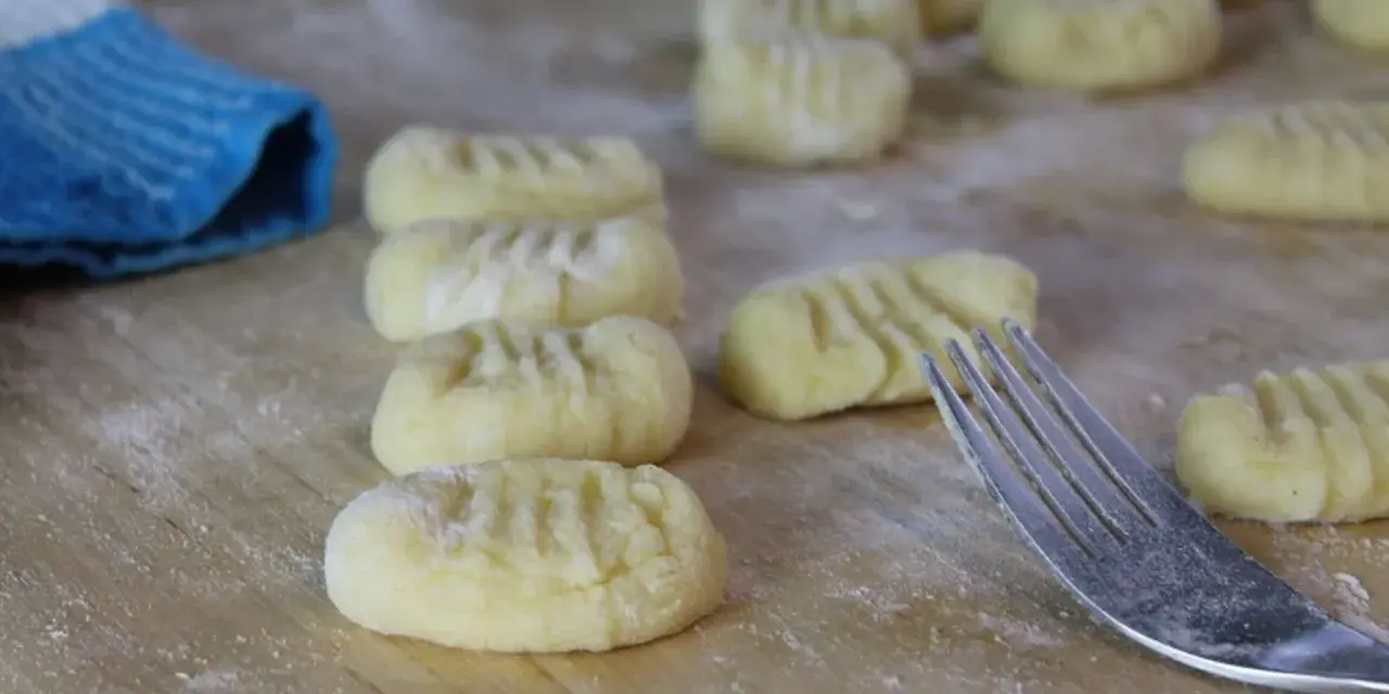 Frisch geformte italienische Gnocchi liegen auf bemehltem Holz. Eine Gabel und ein blauer Lappen sind ebenfalls zu sehen.