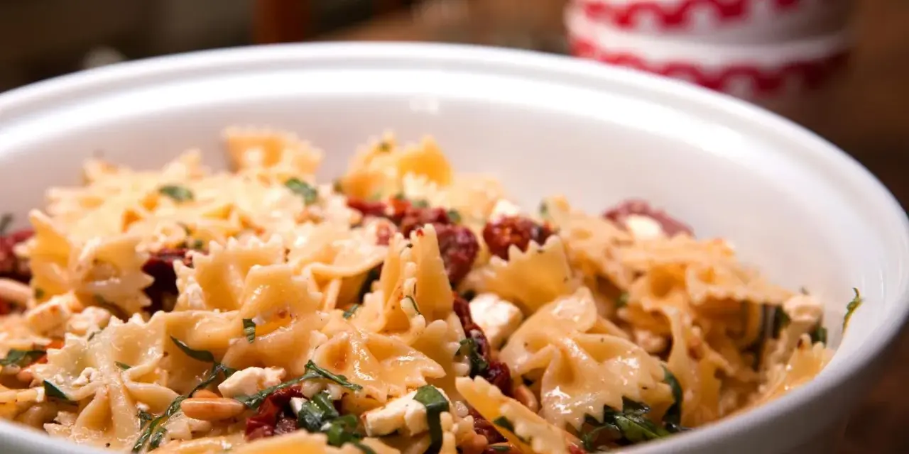 Ein köstlicher italienischer Nudelsalat mit Farfalle, getrockneten Tomaten, Feta und Kräutern in einer weißen Schüssel.