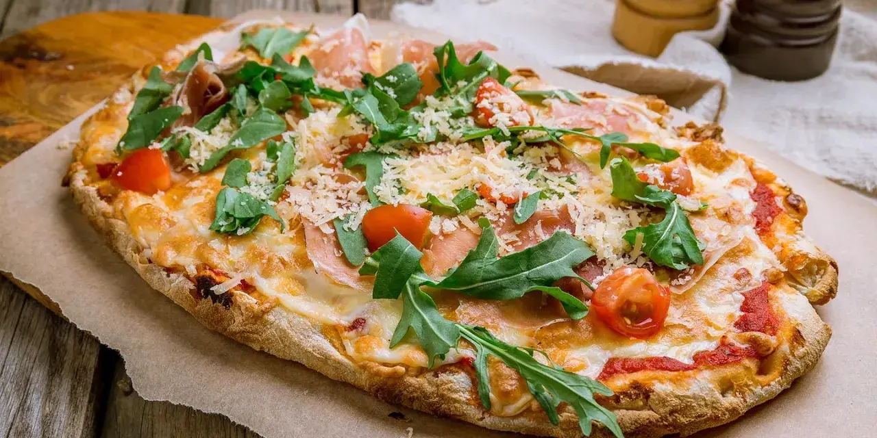 Eine köstliche Pinsa belegt mit Schinken, Rucola, Kirschtomaten und viel Käse auf rustikalem Holzuntergrund.