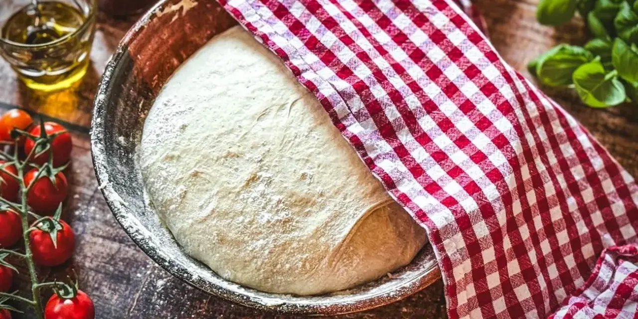 Frischer Pizzateig ruht in einer Schüssel, bedeckt mit einem rot-weiß karierten Tuch. Daneben liegen Kirschtomaten und ein Glas Olivenöl – die perfekten pizzateig zutaten.