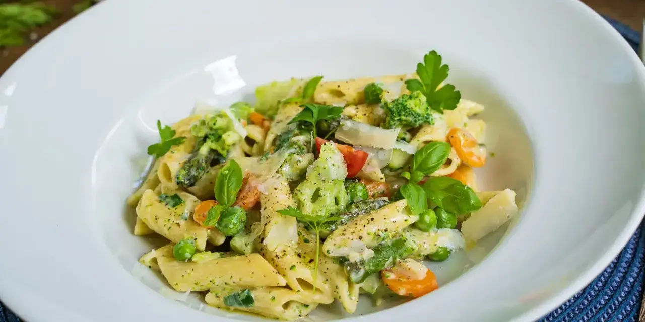 Penne mit cremigem Pesto, Spargel, Erbsen, Brokkoli und Karotten, garniert mit Parmesan und frischen Kräutern.