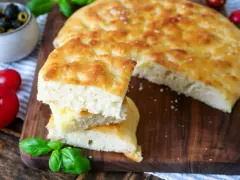 Frisch gebackener focaccia teig, aufgeschnitten und gestapelt, mit Tomaten und Oliven im Hintergrund.