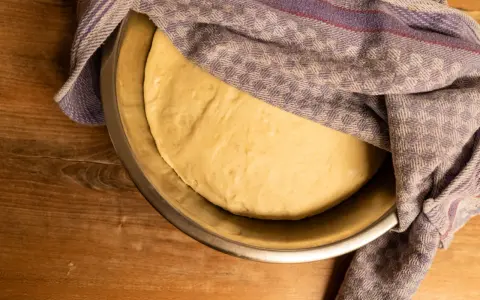 Frischer pizzateig für 4 Personen ruht in einer Schüssel unter einem karierten Tuch auf einem Holztisch.