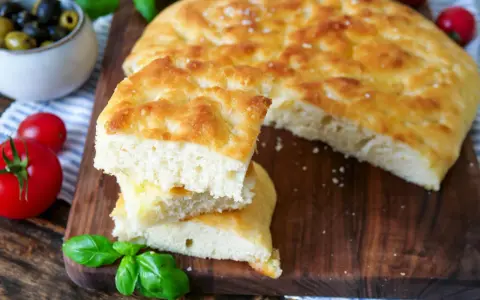 Frisch gebackener focaccia teig, aufgeschnitten und gestapelt, mit Tomaten und Oliven im Hintergrund.