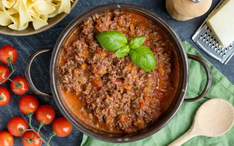 Herzhaftes ragù alla bolognese in einem Topf, garniert mit Basilikum. Daneben frische Tomaten, Bandnudeln und Parmesan.